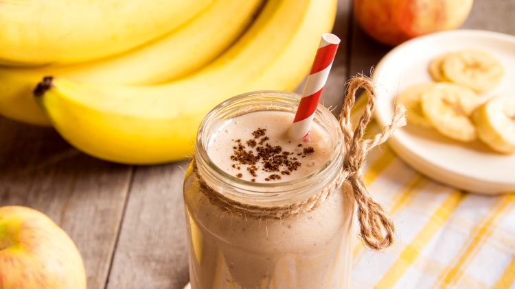 Ein Glas Bananen-Smoothie mit rotem Papierstrohhalm, dekoriert mit Pulver, daneben Bananen, ein Apfel und Bananenscheiben.