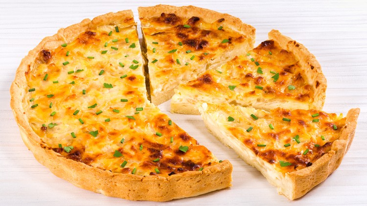 Goldbraun gebackene Quiche mit Käse, Zwiebeln und Schnittlauch belegt, auf weißem Holzuntergrund, mehrere Stücke herausgeschnitten.
