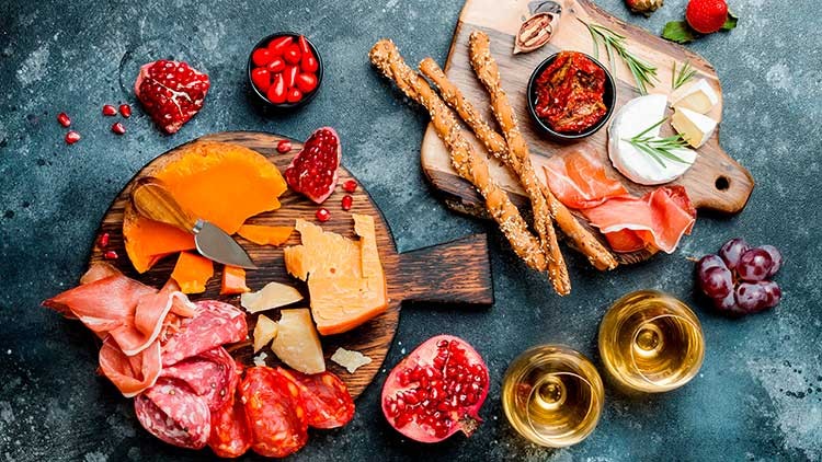 Verschiedene Käsesorten, Wurst, Brotstangen, Obst, Granatapfelkerne und zwei gefüllte Weingläser auf Holzbrett und Tisch.