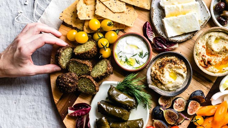 Holzplatte mit Falafelbällchen, Oliven, Brot, Käse, Hummus, gefüllten Weinblättern, Gemüse, Feigen und zwei Händen am Rand.