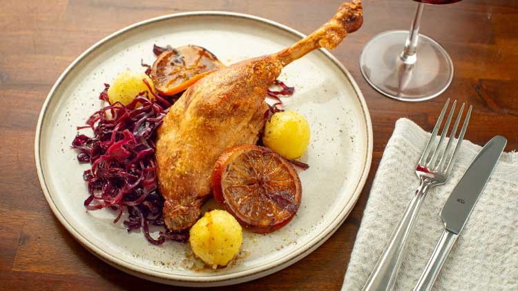 Eine knusprige Gänsekeule mit Rotkohl, Kartoffelklößen und Orangenscheiben liegt auf einem weißen Teller, daneben Besteck.