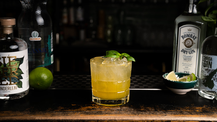 Ein Cocktail mit gelbem Getränk, Eiswürfeln und Basilikum-Dekoration steht auf einer dunklen Bar mit Limette, Flaschen und Schale.