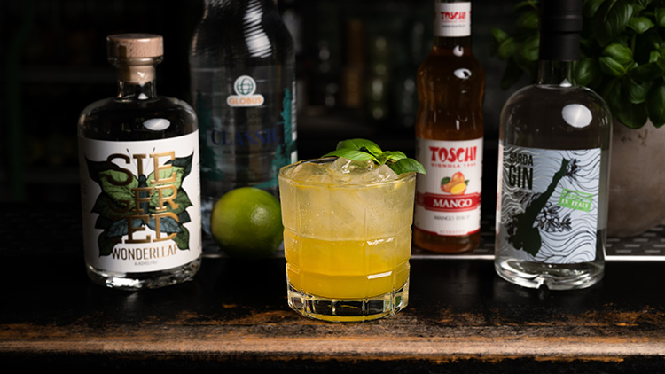 Ein Cocktail mit Basilikum und Eiswürfeln steht auf einer Theke, umgeben von Ginflaschen, Limonade, Mango-Sirup und einer Limette.