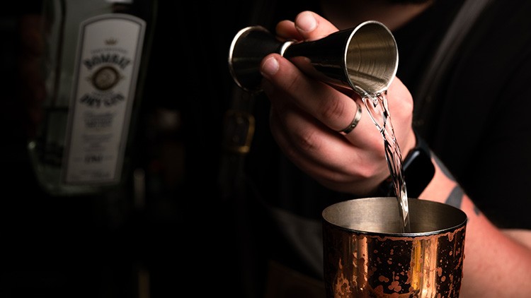 Eine Hand gießt Flüssigkeit aus einem Messbecher in einen kupferfarbenen Becher, daneben steht eine Flasche Bombay Dry Gin.