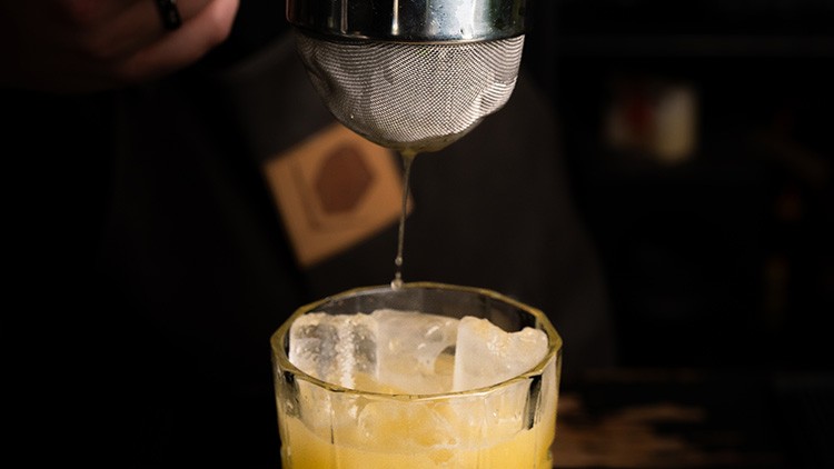 Ein Sieb tropft gelbe Flüssigkeit in ein Glas mit Eiswürfeln vor dunklem Hintergrund.