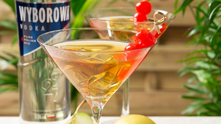 Ein Cocktailglas mit gelblichem Getränk und roten Kirschen steht vor einer Wodkaflasche, einer Zitrone und einem Apfel.