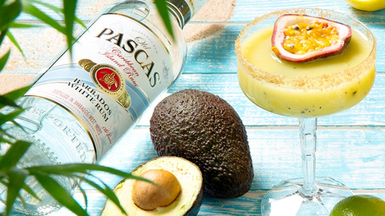 Flasche weißer Rum der Marke Pascas, Cocktailglas mit Passionsfrucht, Avocado, Limetten und Zitronen auf blauem Holztisch.