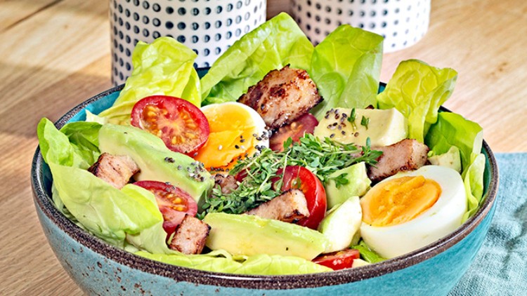 Farbschale mit Salat aus Kopfsalat, Tomaten, Avocado, Eiern, Croûtons und Kresse neben gemustertem Krug und Zuckerbehälter.