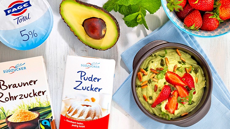 Schale mit grünem Avocadodessert, garniert mit Erdbeeren und Minze, umgeben von Zutaten wie Joghurt, Zucker, Avocado und Erdbeeren.