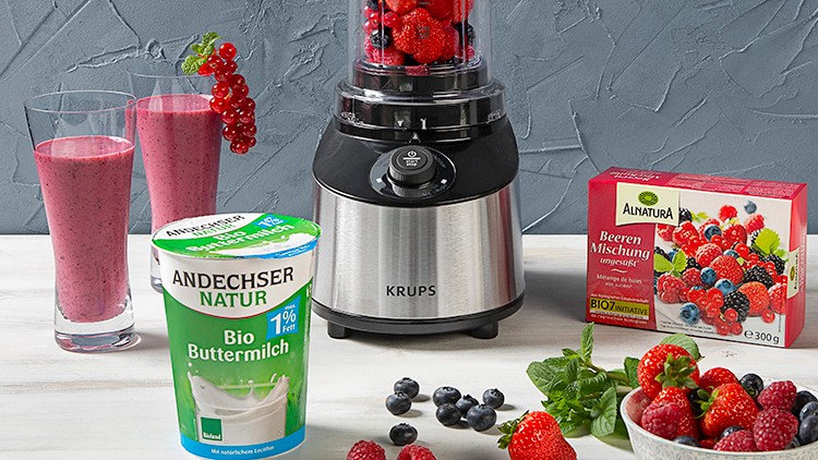 Ein Mixer mit roten Beeren, zwei Gläser roter Smoothie, Bio-Buttermilch, Beerenmischung, Flohsamenschalen und frische Beeren.