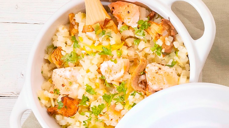 Weiße Auflaufform mit Griffen, gefüllt mit gebratenem Lachs, Blumenkohlreis, Pfifferlingen und frischen Kräutern, Holzlöffel liegt darin.