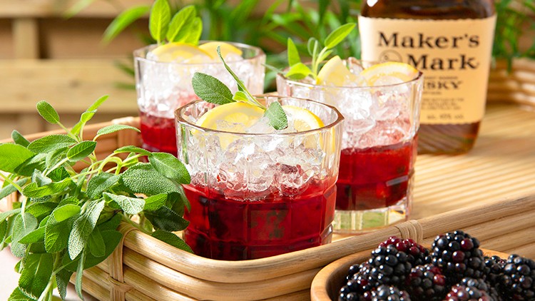 Drei Gläser mit rotem Getränk, Eiswürfeln, Zitronenscheiben und Salbeiblättern stehen auf einem Tablett neben Brombeeren.