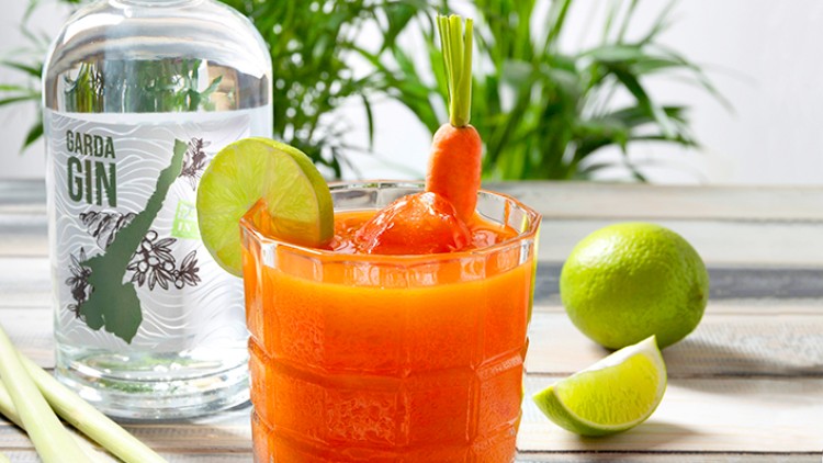Ein Glas mit orangefarbenem Cocktail, Karottengarnitur und Limettenscheibe, daneben eine Flasche Garda Gin und Limetten.