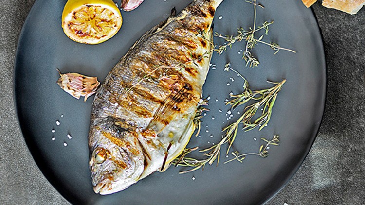 Gegrillter ganzer Fisch auf einem dunklen Teller mit Kräutern, Knoblauch, Zitrone, Salz, Brot, Stoffserviette und zwei Gläsern Weißwein.