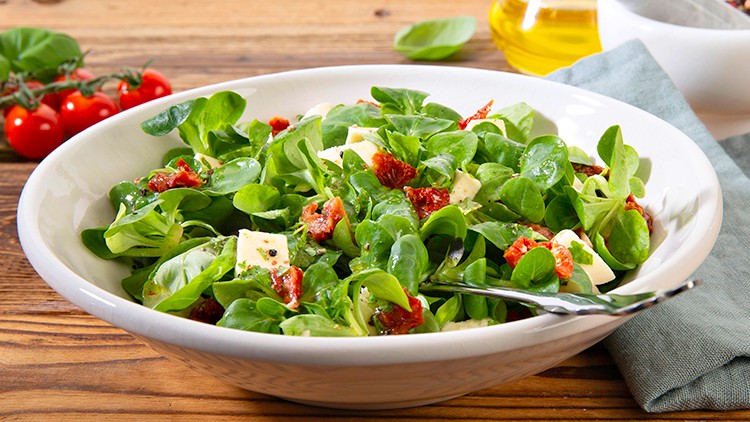 Schale mit grünem Feldsalat, getrockneten Tomaten, Käsewürfeln und schwarzem Pfeffer, daneben Ölflasche und Mörser.