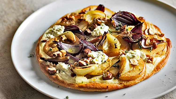 Runde Tarte mit roten Zwiebeln, Birnenscheiben, Käse und Nüssen auf weißem Teller mit Weinglas im Hintergrund.