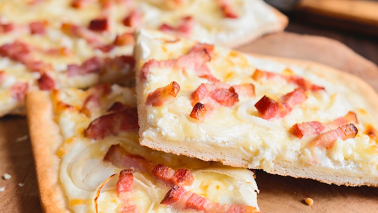 Knuspriger Flammkuchen mit Speckwürfeln und Zwiebelringen auf einem rustikalen Holzbrett, im Hintergrund frische Zutaten.