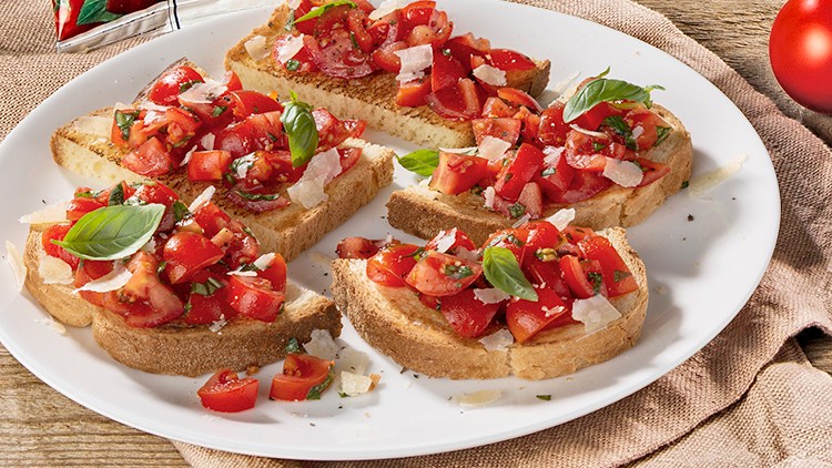 Fünf Scheiben knuspriges Brot mit frischen Tomaten, Basilikum und Käseraspeln auf einem weißen Teller, daneben liegen Tomaten.