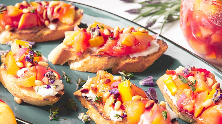 Sechs mit Frischkäse und Pfirsichchutney belegte Brotscheiben auf grünem Teller neben Pfirsich und Glas mit Chutney.