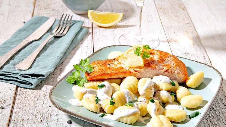 Gekochte Gnocchi mit heller Soße und gebratenem Lachs auf einem Teller, daneben Zitronenscheiben, Besteck und ein gefülltes Weinglas.
