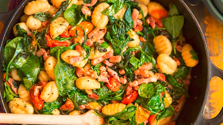 Gnocchi mit Blattspinat, Tomaten und Speckwürfeln in einer schwarzen Pfanne, Holzlöffel liegt darin.