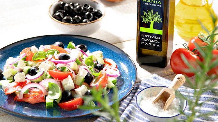 Ein Teller mit griechischem Salat, schwarze Oliven, Brot, Olivenölflasche und Salzschale auf hellem Holztisch.