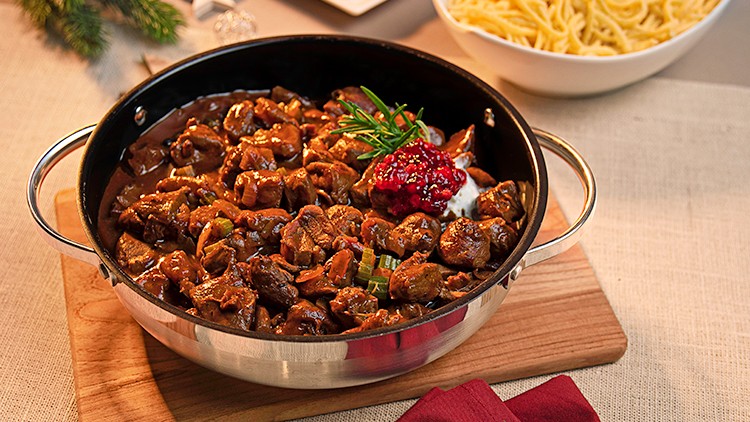 Ein Topf mit Wildragout, dekoriert mit Rosmarin, Sahne und Preiselbeeren, daneben Nudeln und Möhren auf einer gedeckten Tafel.