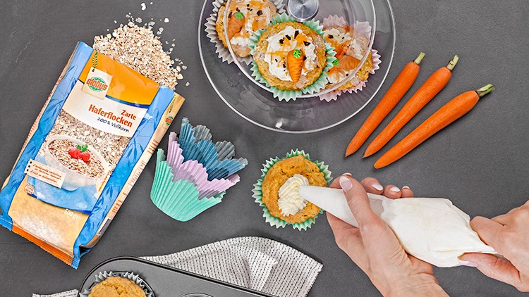 Person dekoriert Muffin mit Spritztüte auf grauer Oberfläche, daneben Haferflocken, bunte Förmchen, Marzipanmöhren und Verpackung.