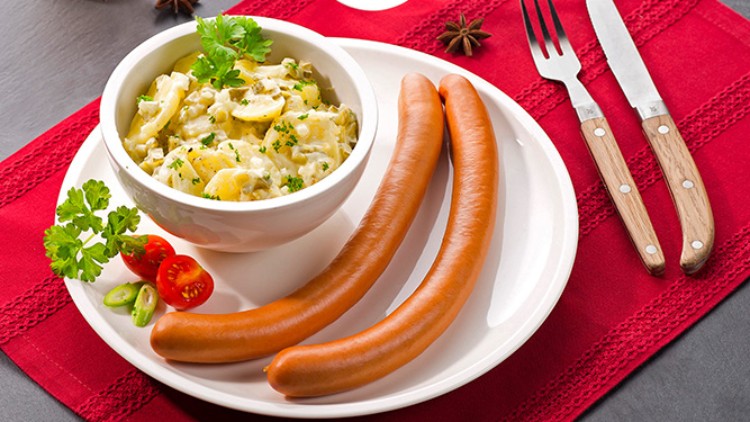 Ein Teller mit zwei Würstchen, Kartoffelsalat und Dekoration aus Kräutern und Gemüse auf roter Tischdecke mit Besteck und Getränken.