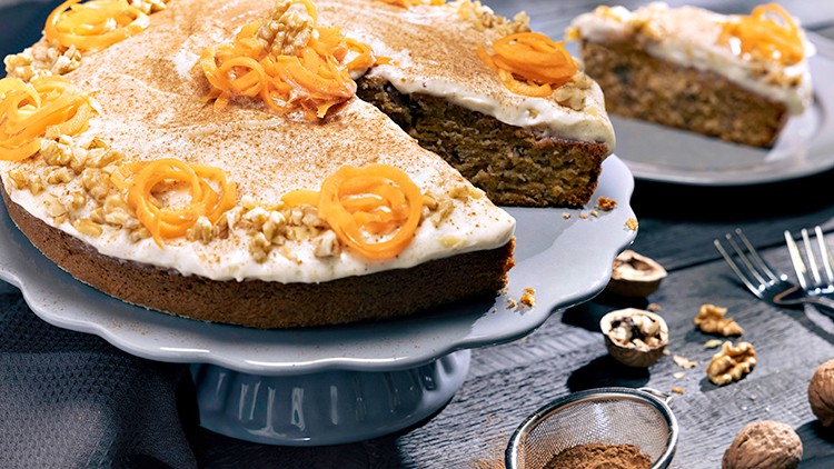 Runder Karottenkuchen mit weißem Topping, Nüssen und Deko aus Karottenstreifen auf einer Tortenplatte, daneben ein angeschnittenes Stück.