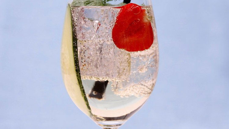 Weinglas mit klarem Getränk, Eiswürfeln, Limettenscheibe, Erdbeere, Minze und schwarzem Strohhalm vor blauem Hintergrund.