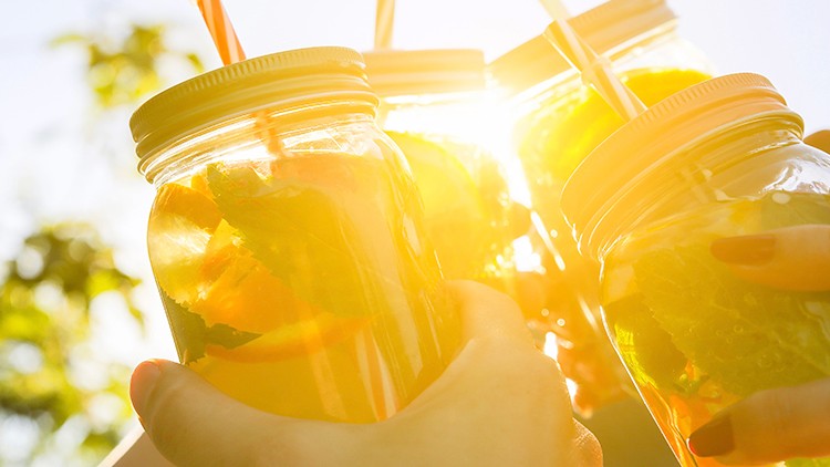 Vier Gläser mit gelbem Getränk, Zitronenscheiben und Minzblättern werden draußen im Sonnenlicht mit Strohhalmen angestoßen.