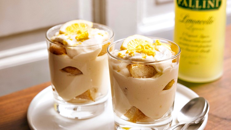 Zwei Gläser mit Dessert, dekoriert mit Zitronenscheiben und geriebener Zitronenschale, stehen auf einem weißen Teller mit zwei Löffeln.