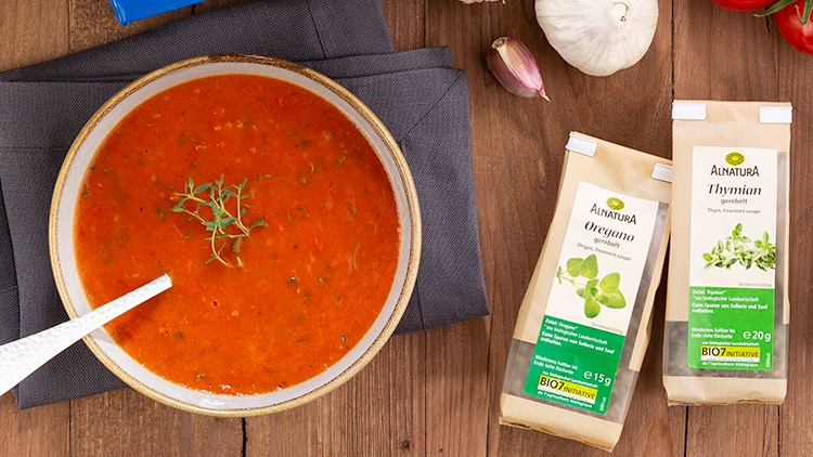 Holzuntergrund mit Thymian, Tomaten, Knoblauch, Alnatura-Produkten, Brot, Essigflasche und Tomatensuppe in einer Schale.