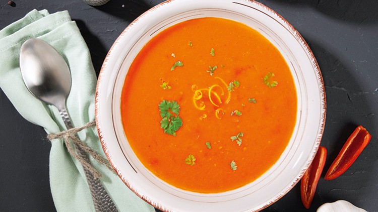 Eine weiße Schale mit orangefarbener Suppe, garniert mit Kräutern und Zitronenzeste, neben einem Löffel und grünem Stoff.