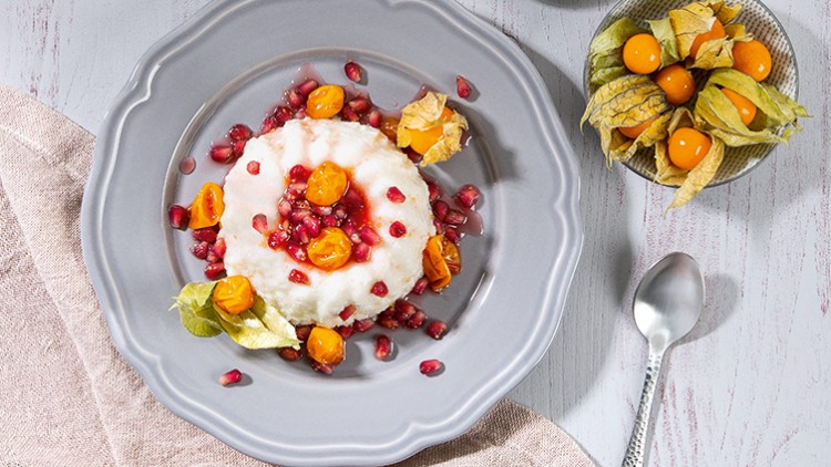 Auf einem grauen Teller liegt eine weiße Süßspeise, dekoriert mit Granatapfelkernen und Physalis, daneben eine kleine Schale und ein Löffel.