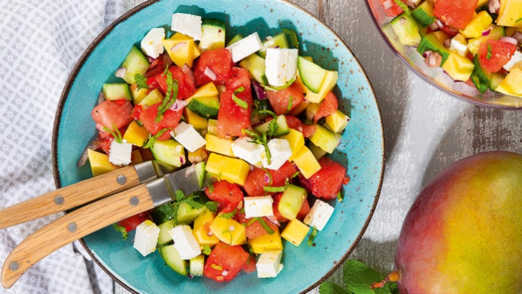 Ein bunter Salat aus Wassermelone, Mango, Gurke, Feta und Kräutern in einer blauen Schale mit zwei Holzgriffmessern.