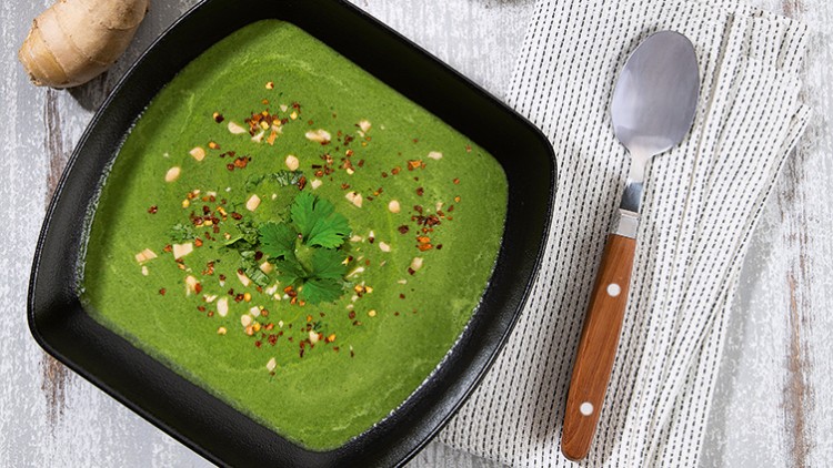 Grüne Suppe in schwarzer Schale mit Kräutern und Nüssen garniert, daneben Löffel auf gestreiftem weißen Stoff.