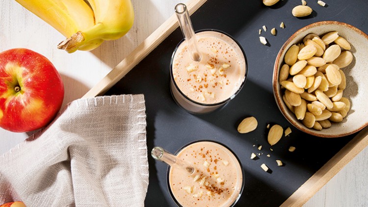 Zwei Gläser Smoothie mit Glasstrohhalmen, daneben ein roter Apfel, Bananen, Mandeln und ein beiges Tuch auf einem schwarzen Tablett.
