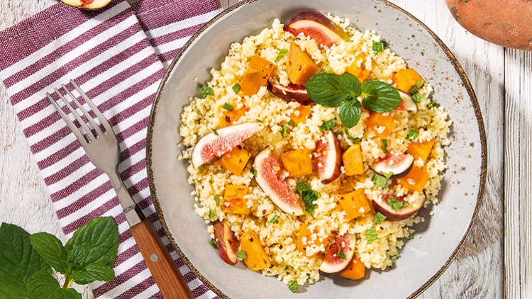 Ein Teller mit Couscous, Stücken von Feigen und Kürbis sowie frischer Minze, daneben eine gestreifte Serviette und eine Gabel.
