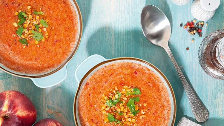 Zwei Schüsseln mit tomatenroter Suppe, garniert mit gehackten Nüssen und Kräutern, mit Löffel, Gewürzen und Pfirsichen auf Holztisch.