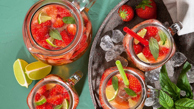 Vier Gläser mit rotem Getränk, Erdbeeren, Limettenstücken und Minzblättern, dazu Eiswürfel und Deko aus frischen Zutaten.