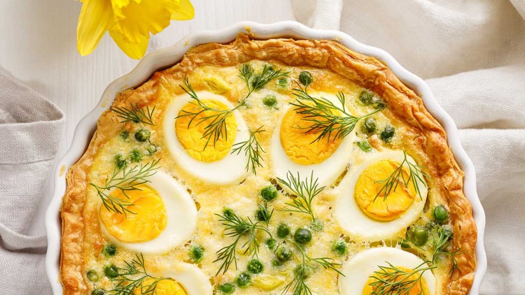 Quiche mit halben gekochten Eiern, grünen Erbsen und Dill in weißer Form, daneben Ostereier und gelbe Narzissen.