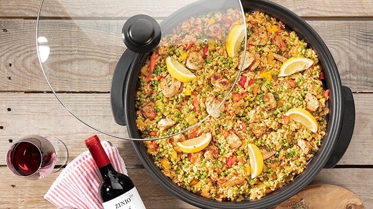 Runde Pfanne mit Paella aus gelbem Reis, Gemüse, Hähnchen und Zitronenspalten, daneben Rotweinflasche und Glas auf Holztisch.