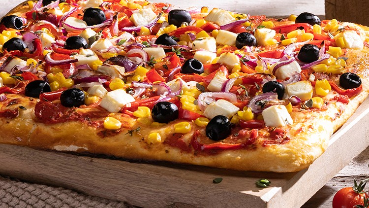 Rechteckige Pizza mit Oliven, Mais, roten Zwiebeln, Paprika und Käsewürfeln auf einem Holzbrett, dekoriert mit Kräutern und Kirschtomaten.