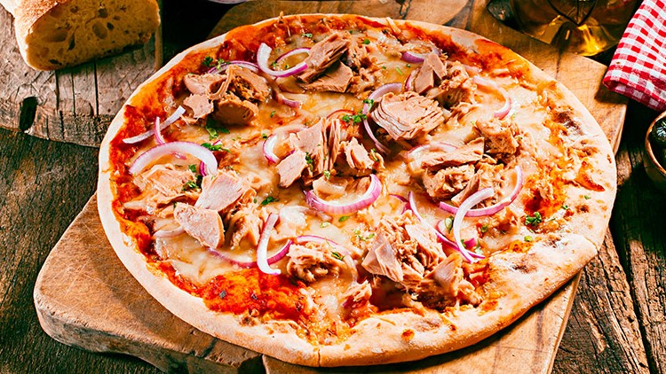 Pizza mit Thunfisch, roten Zwiebelringen und Käse auf Holzbrett, daneben Brot, Oliven, Ölflasche und rot-weiß kariertes Tuch.