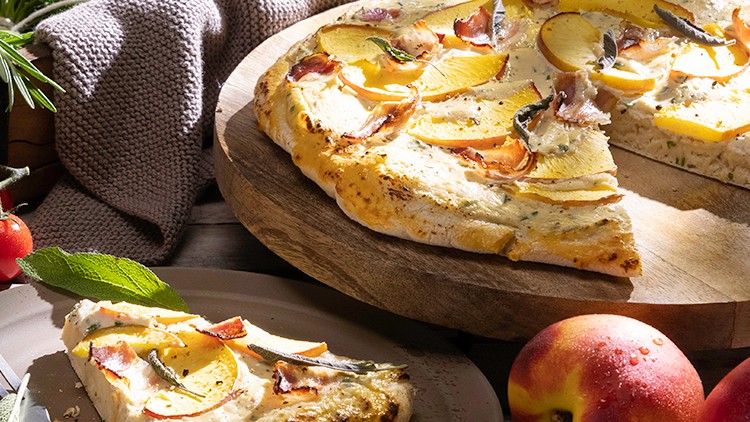Eine goldbraune Flammkuchen-Pizza mit Apfelscheiben und Kräutern auf Holzbrett, daneben frische Zutaten wie Tomaten und Pfirsiche.