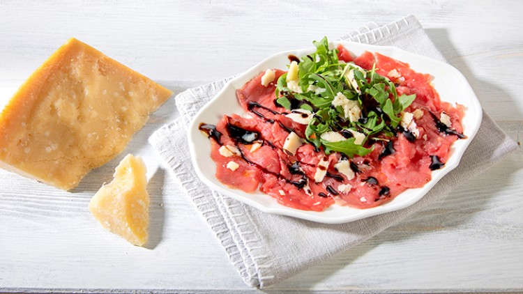 Eine weiße Schale mit Carpaccio, Rucola, Parmesan und Balsamico steht neben einem großen Stück Parmesan auf einem hellen Untergrund.