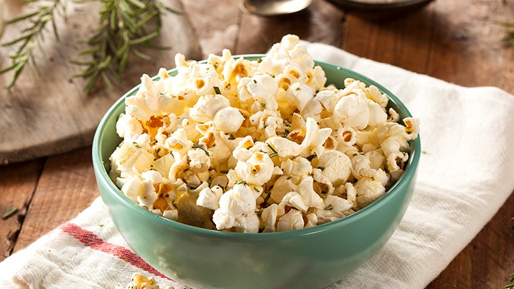Frisch zubereitetes Popcorn in einer grünen Schale auf einem weißen Tuch, daneben Knoblauch, Rosmarin und geriebener Käse.