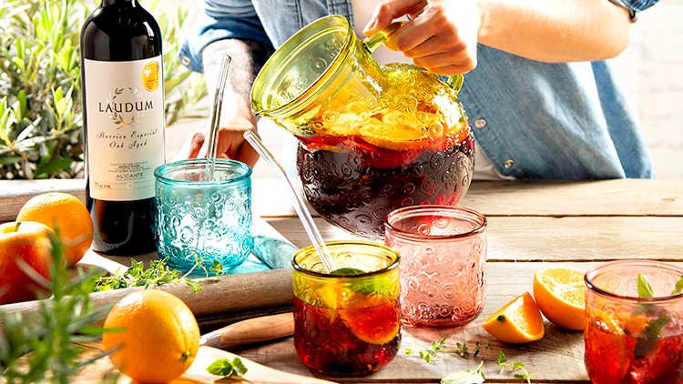 Person gießt roten Fruchtmix aus einer Glaskaraffe in ein buntes Glas, auf einem Holztisch mit Orangen, Kräutern und einer Weinflasche.